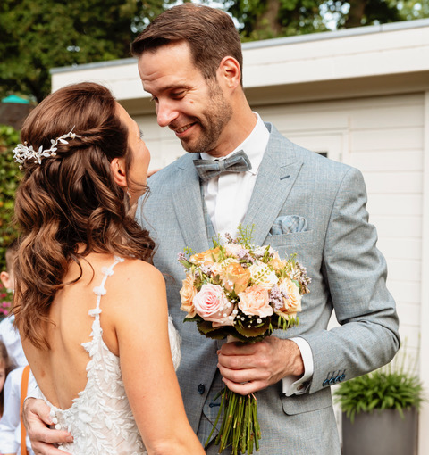 real bride sanneke – romantische A-lijn trouwjurk – bruid en bruidegom kijken elkaar liefdevol aan; de bruidegom houdt het bruidsboeket vast terwijl ze dicht bij elkaar staan